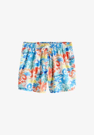 Uomo in piedi vicino alla piscina che indossa pantaloncini da bagno colorati con stampe tropicali di palme, barche e creature marine.