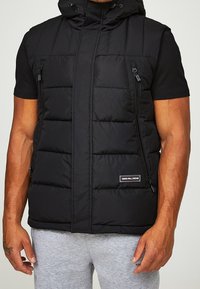 Svart vadderad väst med huva, utrustad med fickor med dragkedja och en logotypapplikation. Tillverkad av texturerat tyg, med en kortad design och utan ärmar.