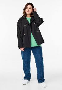 Giacca nera doppiopetto con bottoni argentati, indossata sopra un maglione verde e jeans blu, presentata da un modello con capelli mossi.