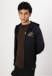 Črna zip-up kapuca z sprednjimi žepi, ki ima grafični dizajn in napis "FORT WORTH, TEXAS." Običajna je nad rjavim majico. Minimalističen stil.