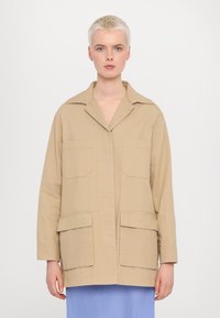 Beige bavlněná pracovní bunda s límcem, dvěma náprsními kapsami a dvěma spodními kapsami. Má uvolněný střih a šití.