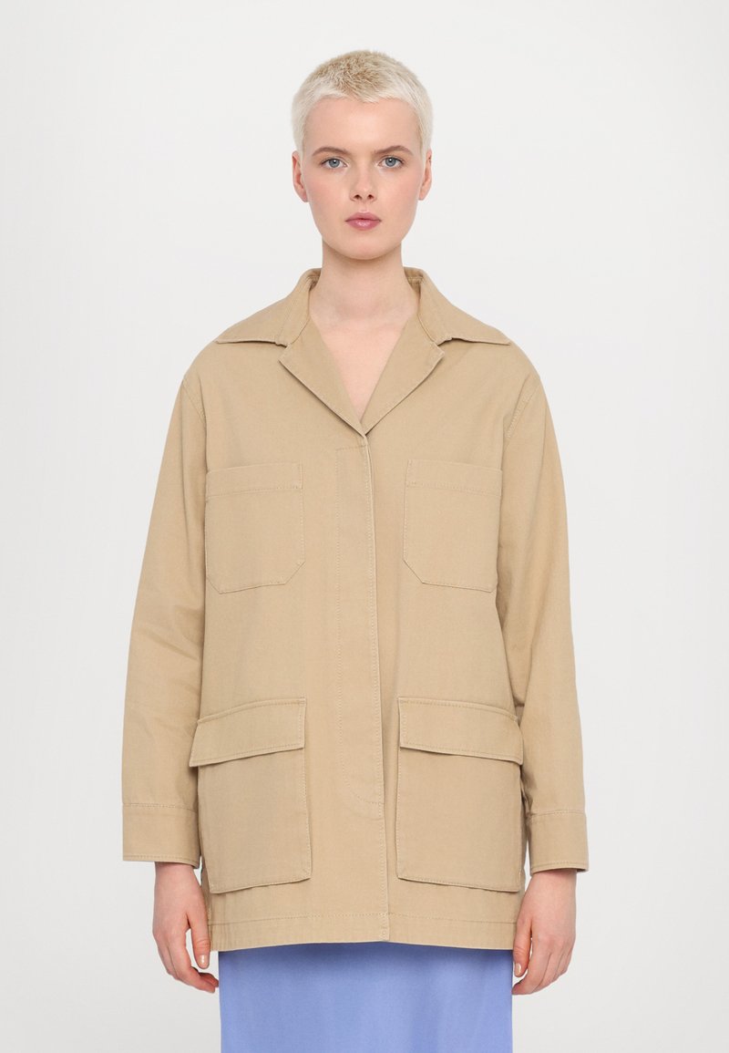 Beige bavlněná pracovní bunda s límcem, dvěma náprsními kapsami a dvěma spodními kapsami. Má uvolněný střih a šití.