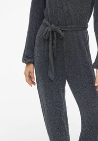 Juodas šviesuotas kombinezonas su diržo juosmeniu, ilgomis rankovėmis ir tekstūruotu blizgesiu, pasižymintis laisvu siluetu ir minkšta medžiaga.