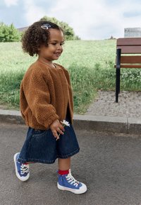 Brun teddyfrakke, denimnederdel og blå høje sneakers med røde detaljer. Barn holder en blomst og går på en brolagt sti nær græs.