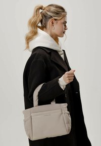 Beige vuorattu käsilaukku, jossa on kaksi lyhyttä kantokahvaa, ja joka on varustettu sileällä pinnalla ja minimalistisella muotoilulla. Käytössä henkilöllä, joka on pukeutunut mustaan takkiin.