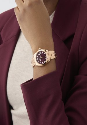 Roségoldene Armbanduhr mit Zifferblatt in rundem Burgunderrot, Metallarmband und einfachen Stundenmarkierungen, die auf einem Handgelenk unter dem Ärmel eines burgunderfarbenen Blazers ruht.