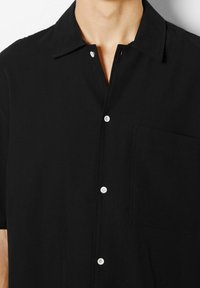 Czarna koszula z krótkim rękawem i ostrym kołnierzykiem, z białymi guzikami i pojedynczą kieszenią na piersi. Materiał wydaje się gładki o matowym wykończeniu.