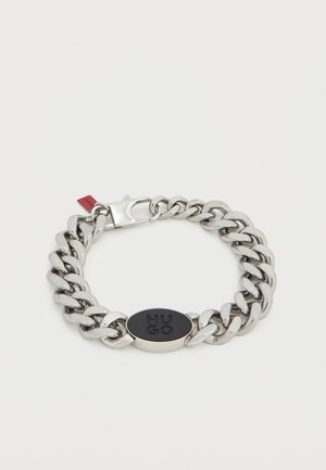 Metalen armband met grote, in elkaar grijpende schakels, gepolijste zilveren afwerking, ovale zwarte logo-accent en beveiligde sluiting.