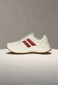 Baskets en cuir blanc avec des rayures rouges, une semelle couleur tan et le logo "BALLY" doré sur le côté, présentées sur une surface gris clair.