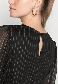 Vestido preto com riscas verticais douradas. Mangas bufantes transparentes e abertura em forma de buraco-chave nas costas, segurada com um pequeno botão.