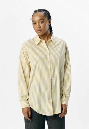 Chemise à rayures verticales jaunes et blanches à manches longues avec un devant à bouton et un col pointu, associée à un pantalon gris foncé.