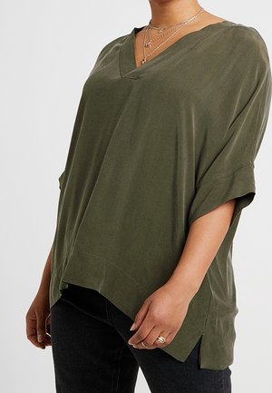 Persona vistiendo una blusa holgada verde oliva con cuello en V y mangas cortas remangadas, pantalones negros, y accesorios con collares dorados en capas y anillos.