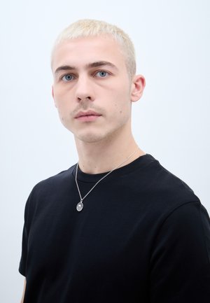 Fekete, rövid ujjú póló, ezüst színű medál nyaklánccal, textúrált, csíkozott ovális mintával; minimális háttér.