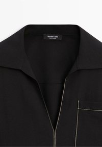 Chemise noire avec col ouvert, poche avant visible et surpiqûres blanches contrastantes le long de la fermeture éclair et des bords de la poche.