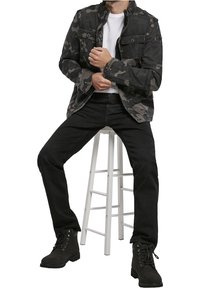 Svart kamouflagejacka med knäppte fickor, matchad med svarta jeans och svarta snörskor, sittande på en vit träpall.