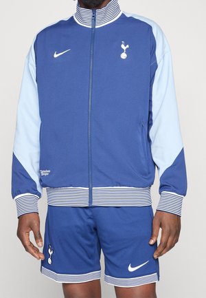 Modèle portant un survêtement bleu et bleu clair de Tottenham Hotspur, avec une veste zippée, des finitions à rayures, l’écusson du club et des logos Nike.