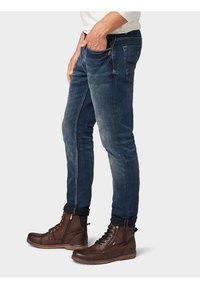 Vaqueros de mezclilla azul oscuro con corte ajustado, que presentan un sutil desgaste y un diseño clásico de cinco bolsillos, combinados con botas marrones de cordones.