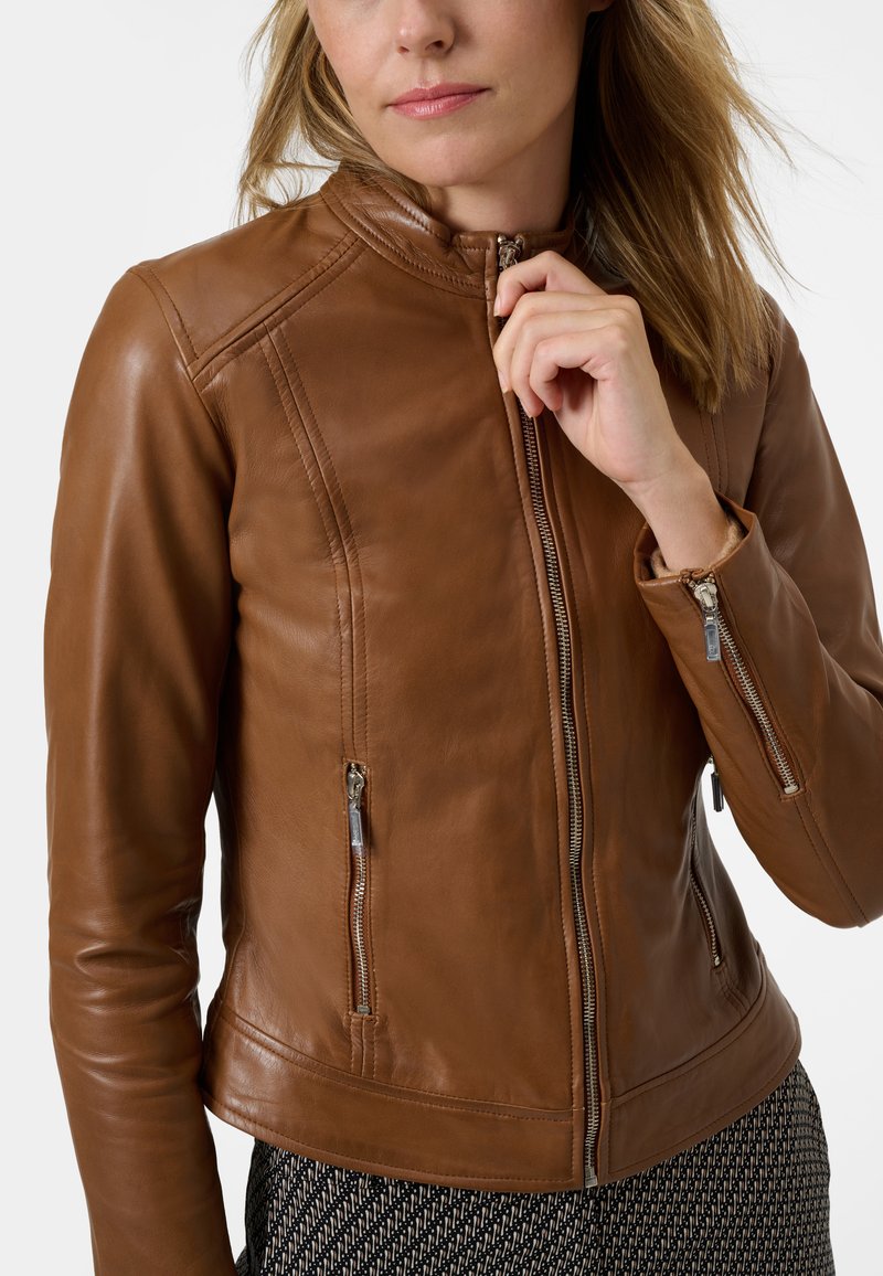 Veste en cuir marron avec un design ajusté, col haut, fermeture éclair à l'avant et deux poches zippées. Présente des accents de couture subtils.