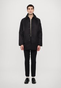 Black, waterproof jacket with a high collar, snap button front, two side pockets, and a smooth texture, paired with slim black pants.
