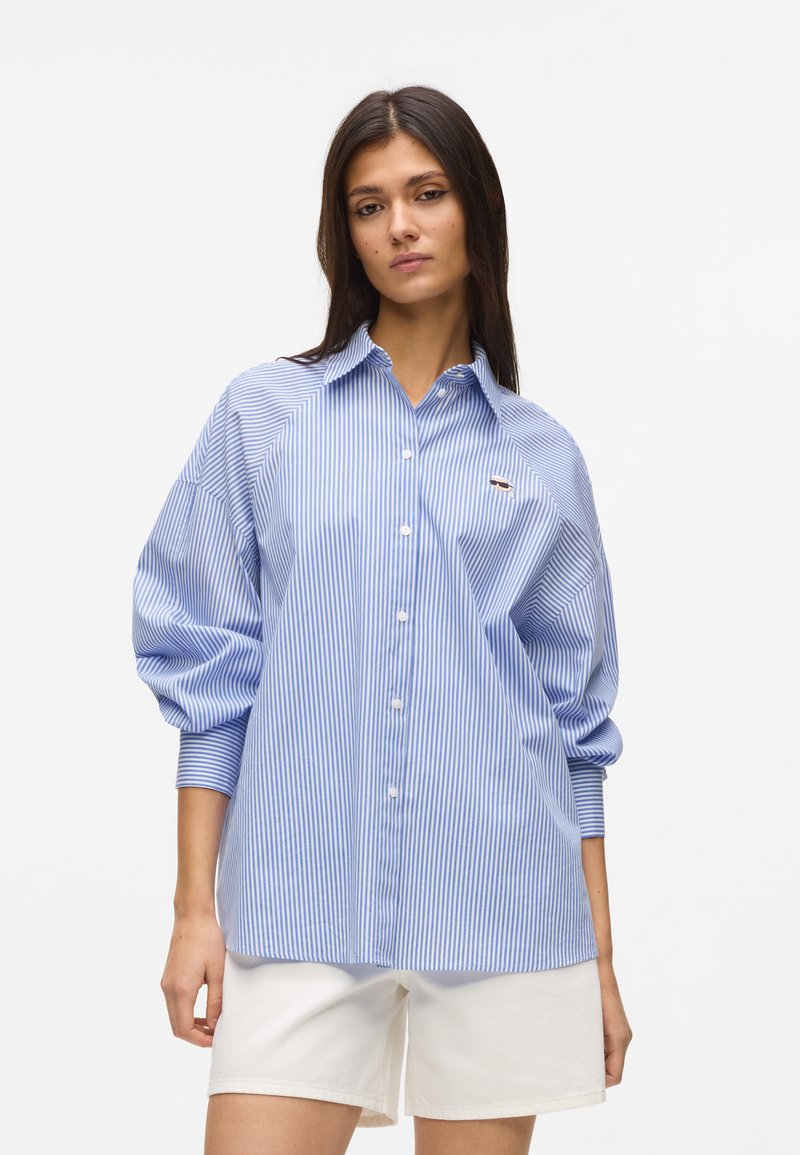 Γυναίκα που φοράει ανοιχτό μπλε και λευκό κάθετο ριγέ πουκάμισο με φαρδιές μανσέτες και λευκά σορτς, στέκεται μπροστά σε απλό φόντο.