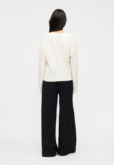 Γυναίκα που στέκεται με την πλάτη, φοράει λευκό πουλόβερ με μακριά μανίκια και φαρδιά μαύρη παντελόνα, πάνω σε απλό φωτεινό φόντο.