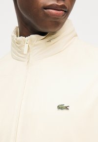 Άνδρας που φοράει μπεζ μπουφάν με φερμουάρ και όρθιο γιακά, με μικρό πράσινο λογότυπο κροκοδείλου στο στήθος.