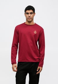 Bordowa koszulka z długim rękawem wykonana z miękkiej bawełny, z haftowanym złotym wzorem po lewej stronie klatki piersiowej i okrągłym dekoltem.