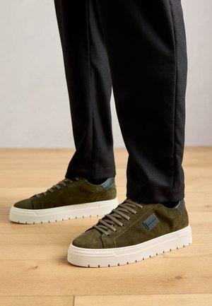 Person wearing olive green suede sneakers with white thick soles and black pants standing on wooden floor.