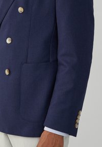 Marinblå dubbelknäppt blazer i texturerad tyg, med gyllene knappar och en enstaka bröstficka. Klassisk skräddarsydd siluett med stiliga manschettdetaljer.