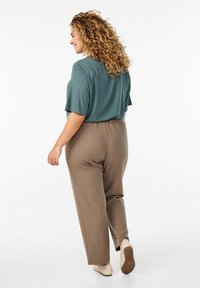 Una persona dai capelli ricci indossa una maglietta oversize di colore teal e pantaloni tapered di un marrone chiaro, con scarpe beige, vista di spalle su uno sfondo bianco.