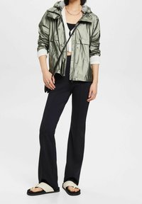 Srebrno-zielona, lekka kurtka przeciwdeszczowa z kapturem, z dwiema przednimi kieszeniami, nałożona na czarny top crop i zestawiona z czarnymi spodniami dzwonami oraz sandałami.