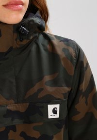 Camouflagejakke i brune og grønne toner, lavet af et glat, vandafvisende materiale, med lynlås og mærkepatch på frontlommen.