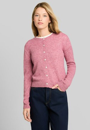Jonge vrouw met een roze gebreid vest met knopen en donkerblauwe spijkerbroek, staand tegen een effen lichtgrijze achtergrond.