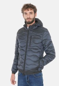 Giacca imbottita nera con motivo a onde, cappuccio e cerniera frontale. Presenta una tasca sul petto con zip e polsini elastici. Indossata con jeans blu.