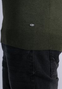 Pull en maille vert foncé avec ourlet côtelé et petit logo ovale métallique de la marque au-dessus de la poche d'un jean en denim noir.