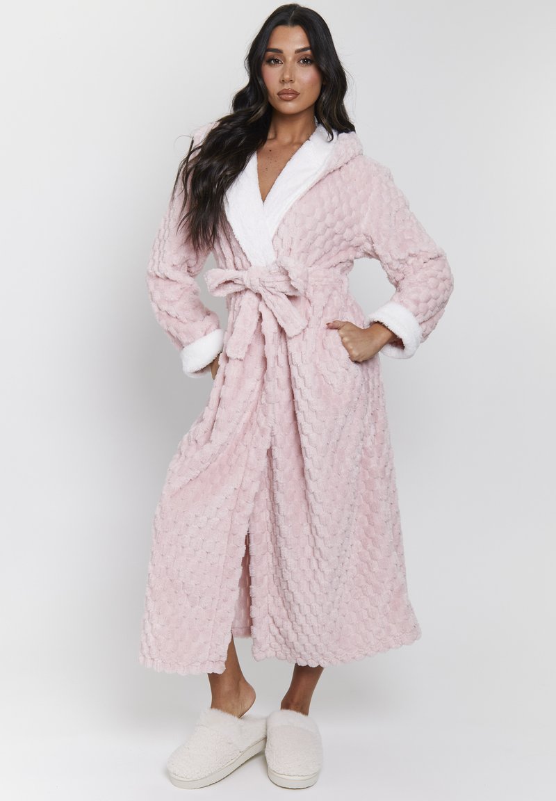 Donna che indossa un accappatoio rosa in tessuto con bordi bianchi, legato in vita, e pantofole bianche pelose, in piedi davanti a uno sfondo semplice.
