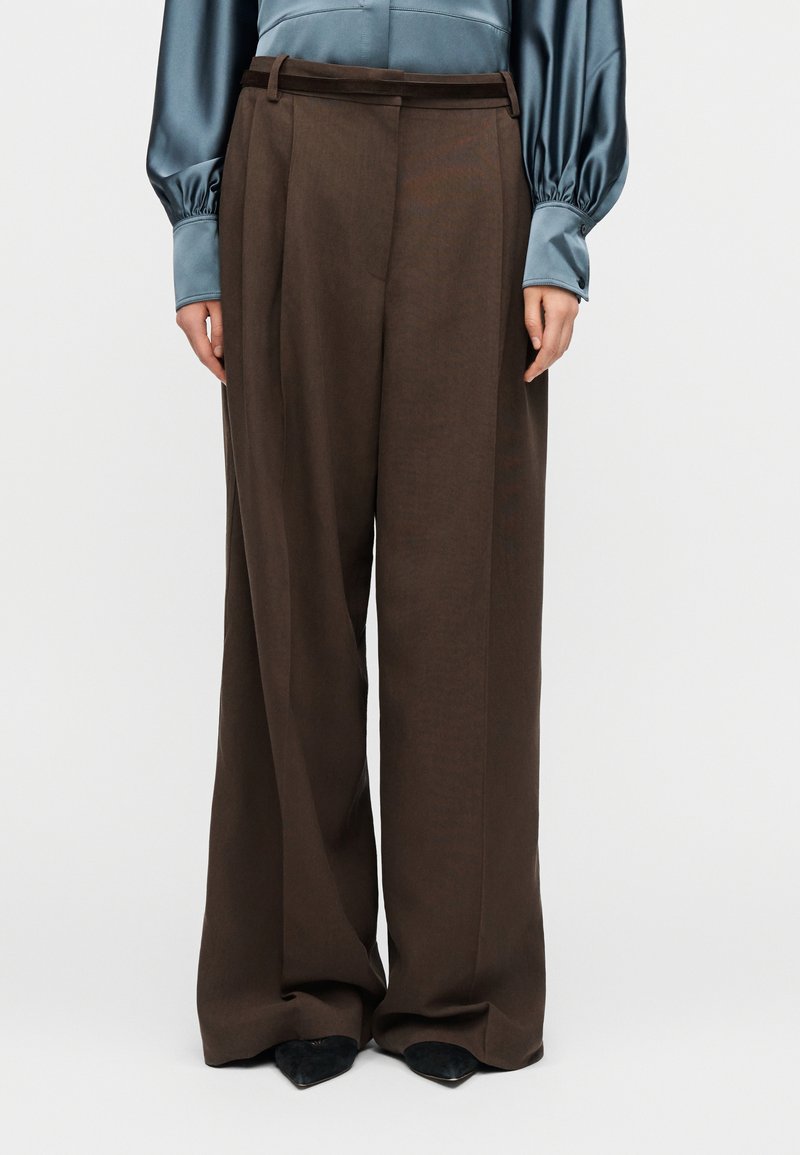 Femme portant un pantalon taille haute, jambes larges, brun foncé, associé à un chemisier bleu en satin à manches longues et des chaussures noires pointues.