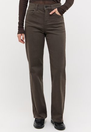 Person, die braune gerade geschnittene Hose, schwarze Schuhe und ein eng anliegendes braunes Langarmoberteil trägt, mit einer Hand in der Tasche.