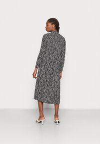 Vestido preto de mangas longas com um pequeno padrão floral branco; possui um comprimento até à metade da panturrilha e um corte reto, combinado com saltos brancos abertos na frente.
