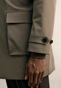 Olivgrüne wasserdichte Jacke mit aufgenähter Tasche, schrägen Ärmelbündchen und schwarzen Knöpfen aus strukturiertem Stoff nahe dem Handgelenk.