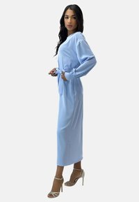 Vestido largo azul claro con mangas largas, cinturón de lazo en la cintura y corte recto. La modelo lleva tacones beige con tiras. Textura de tela lisa.