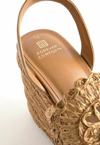 Sandały na wedge w kolorze brązowym z górą z plecionego naturalnego włókna. Zawierają okrągły pleciony akcent oraz wyściełaną wkładkę z detalem w złotym kolorze.