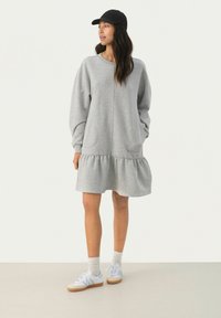 Hellgraues Sweatkleid mit gerafftem Saum, langen Ärmeln und seitlichen Taschen, kombiniert mit einer schwarzen Mütze und weißen Sneakers.