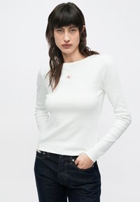 Balts ribots krekls ar garām piedurknēm, uz krūtīm neliels sarkans logo, kopā ar tumši zilām džinsa biksēm. Gluda tekstūra un pieguļošs dizains.