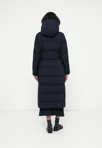 Lang navy pufferjakke med hætte, bælte i taljen og quiltet design. Har en knælang pasform og er parret med en plisseret nederdel.