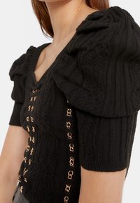 Top nero in maglia con texture, maniche a sbuffo corte, scollo a barca e dettagli in metallo dorato sulla parte anteriore.