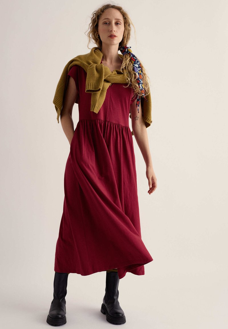 Femme debout dans une robe rouge sans manches, pull jaune noué sur les épaules, bottes noires, cheveux avec foulard à motifs, fond neutre.