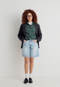 Νεαρή γυναίκα με σγουρά μαλλιά που φορά γυαλιά, ριγέ πράσινο πόλο, μαύρο σακάκι, ανοιχτό τζιν σορτς, λευκές κάλτσες και μαύρα παπούτσια, με τα χέρια στις τσέπες.