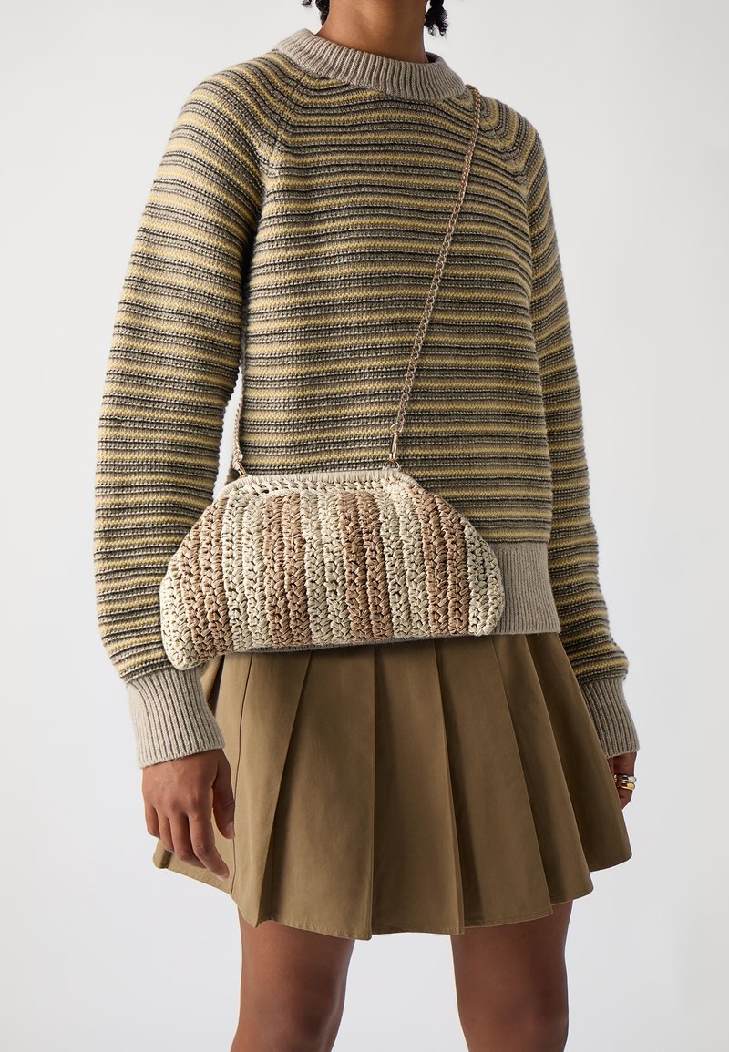 Personne portant un pull en tricot rayé et une jupe plissée kaki, porte un sac à bandoulière tissé beige et marron avec une bandoulière chaîne.