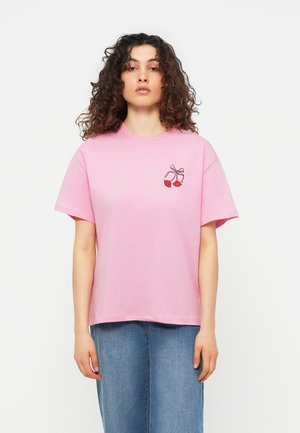 Kvinde med krøllet hår iført en lyserød T-shirt med et motiv af røde læber bundet med et bånd på brystet, kombineret med blå jeans.